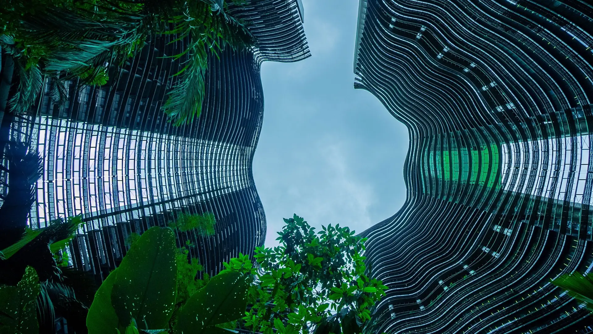 View looking up between two modern, curved high-rise buildings with sleek glass facades, surrounded by lush green plants and trees against a cloudy sky.