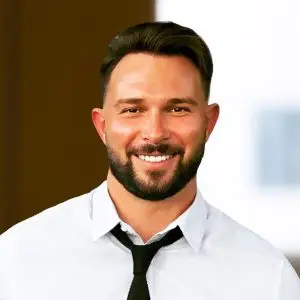 A man with short dark hair and a beard, wearing a light-colored shirt and black tie, smiles confidently while standing indoors with a softly blurred background. Focus keyphrase not set.