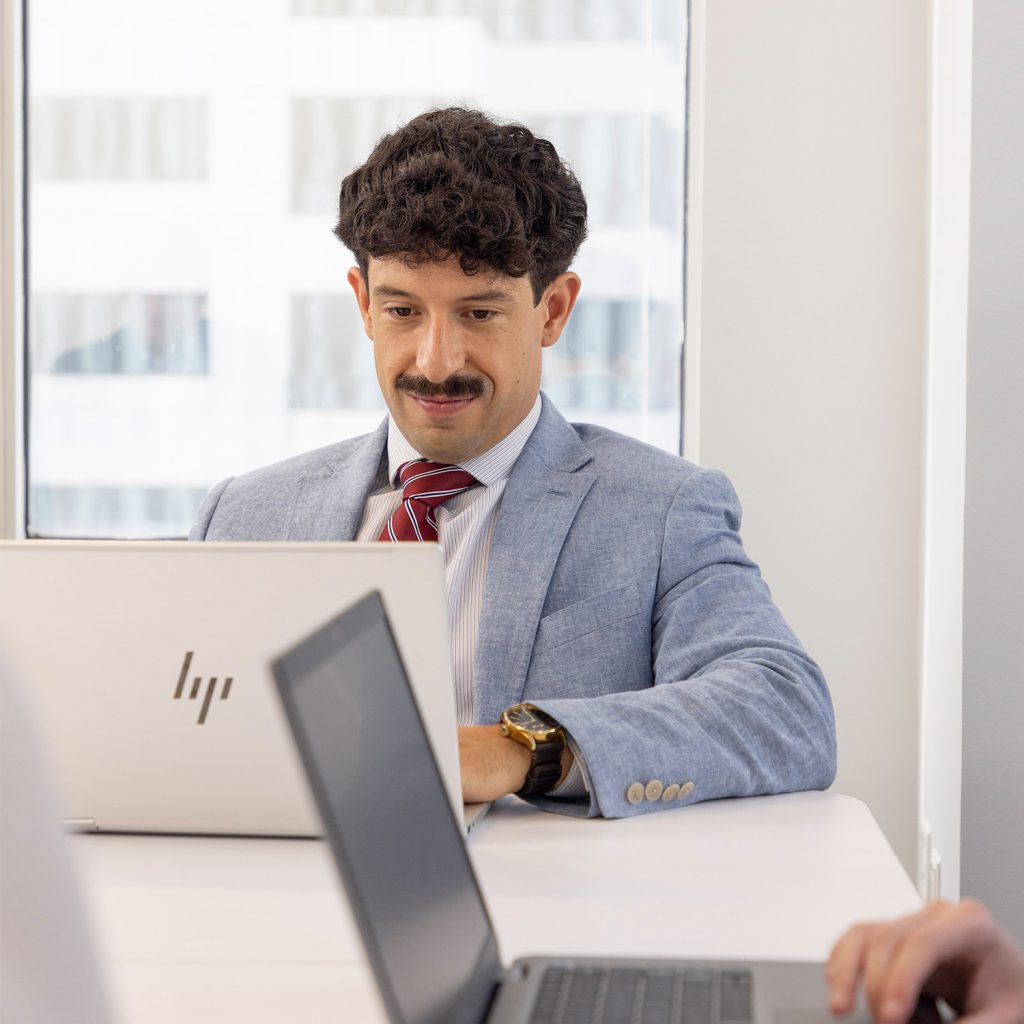 A man in a blue suit and red striped tie works on an HP laptop at a white desk in a modern office, with another person’s laptop visible in the foreground, reflecting the company’s commitment to ISO Certifications.