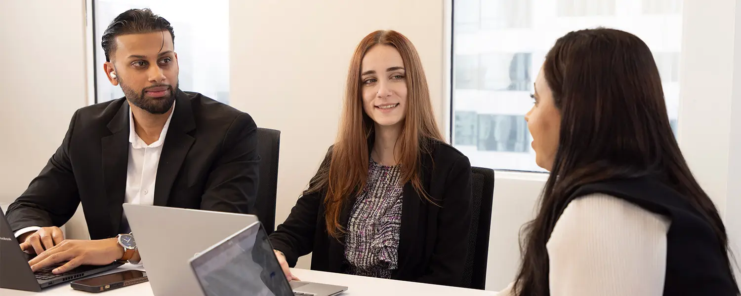 Three people in business attire sit at a conference table with laptops, having a discussion in a modern office setting where the focus keyphrase is not set.
