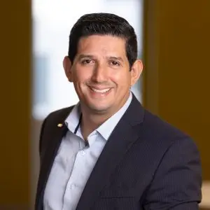 A man wearing a dark suit jacket and light blue shirt smiles at the camera in an indoor setting with a softly blurred background, creating a warm, professional atmosphere.