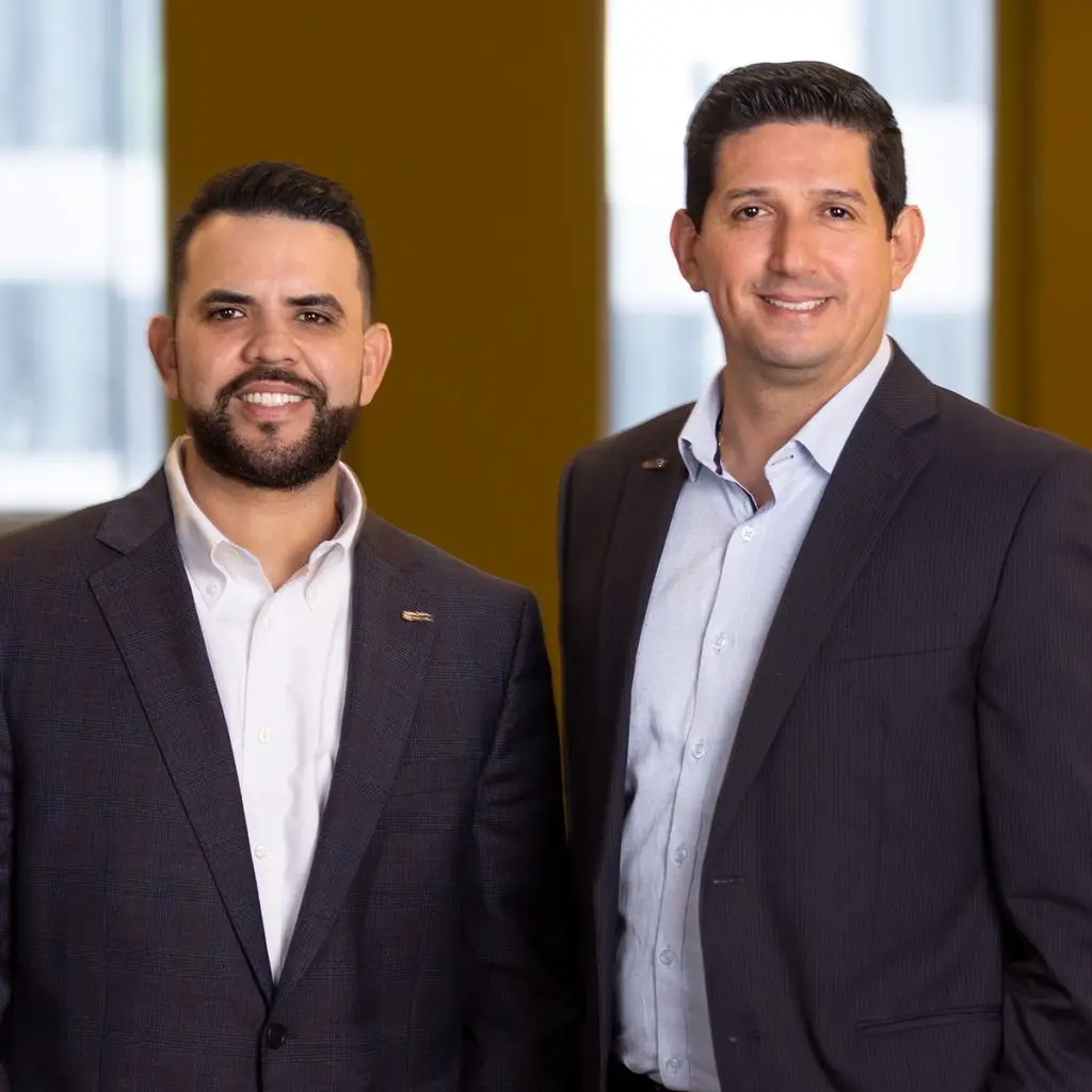 Two men in business suits stand side by side indoors, smiling at the camera with a blurred background of large windows, creating a professional yet friendly atmosphere.