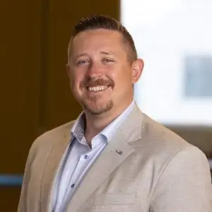 A man with short light brown hair and a trimmed beard, wearing a light beige suit jacket and a light blue shirt, smiling warmly in an indoor setting. Focus keyphrase not set.
