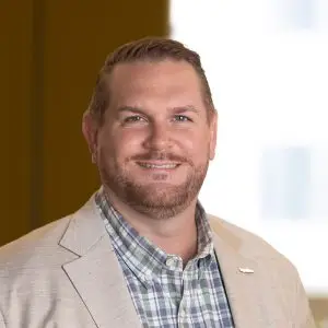A man with short hair and a trimmed beard, wearing a light-colored blazer over a plaid shirt, smiles at the camera, his cheerful expression framed by a softly blurred background. Focus keyphrase not set.