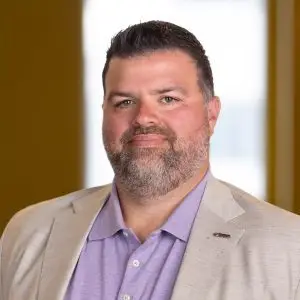 A man with short dark hair and beard, wearing a light-colored blazer and a lavender shirt, stands indoors against a softly blurred background. The focus keyphrase is not set.