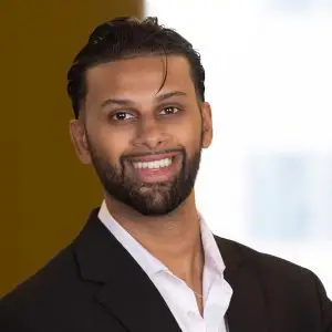 A man with dark hair and a beard, wearing a black suit jacket and white shirt, smiles at the camera in a blurred indoor setting, exuding confidence.