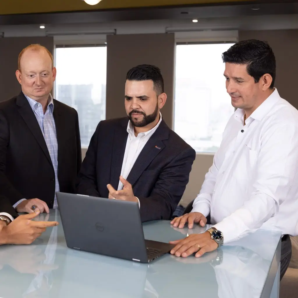 Three men in business attire stand around a glass table, engaged in a discussion as they look intently at a laptop screen in an office setting.