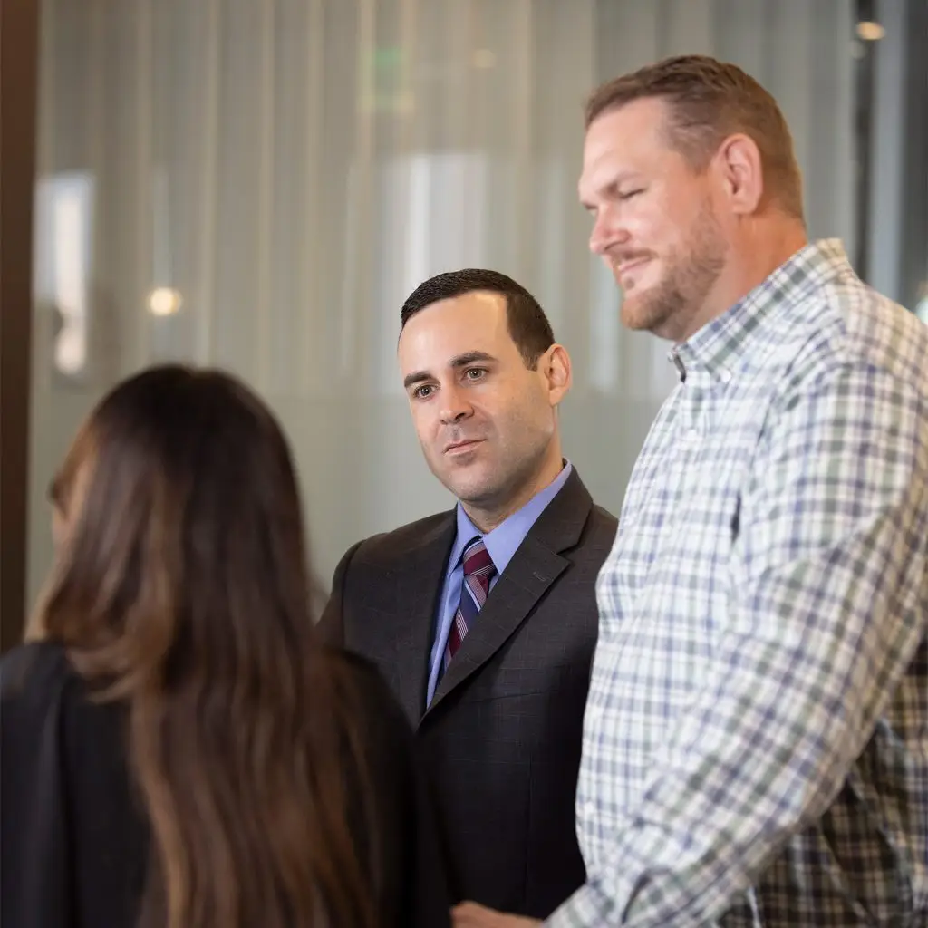 Three people stand indoors having a conversation; one man in a suit faces the camera, another man in a plaid shirt stands beside him, and a woman with long hair—focus keyphrase not set—stands with her back to the camera.
