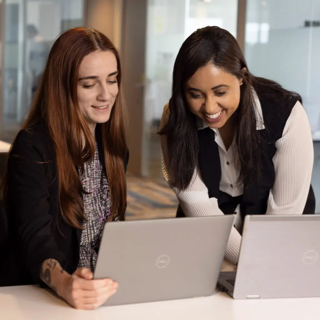 Two women collaborate at a desk, both smiling as they work on their laptops in a bright, modern office setting.