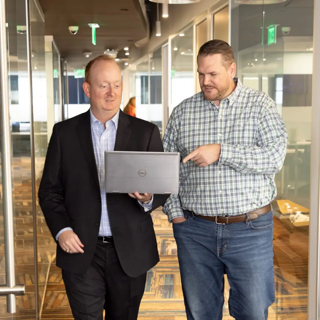 Two men walk down an office hallway; one in a suit holds a laptop while the other, dressed in a plaid shirt and jeans, points at the screen as they discuss their project.