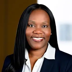 A woman in a dark blazer and white collared shirt smiles at the camera, standing confidently against a blurred office background.