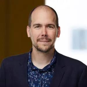 A man with a shaved head and short beard, wearing a dark blazer over a patterned shirt, stands in front of a softly blurred indoor background. Focus keyphrase not set.