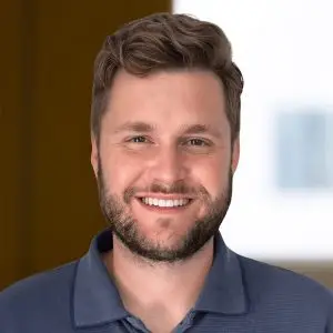 A man with short brown hair and a beard is smiling at the camera, wearing a blue collared shirt, with the focus keyphrase not set against a softly blurred background.