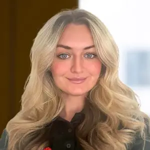 A woman with long blonde hair and blue eyes, wearing a black collared shirt, stands in front of a softly blurred indoor background.