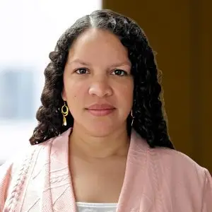 A woman with curly hair, wearing gold hoop earrings and a pale pink textured jacket, stands indoors with a neutral expression and stylish confidence.