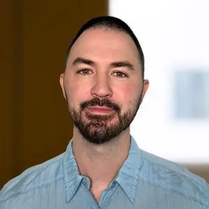 A man with short dark hair and a beard, wearing a light blue collared shirt, stands in front of a softly blurred indoor background—focus keyphrase not set.