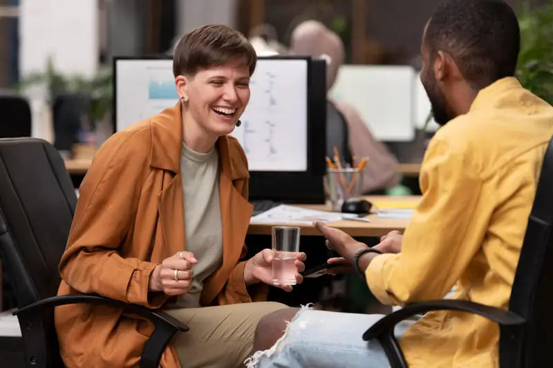 A person holding a glass of water is laughing while talking with another colleague in an office setting, surrounded by computers and various office supplies.