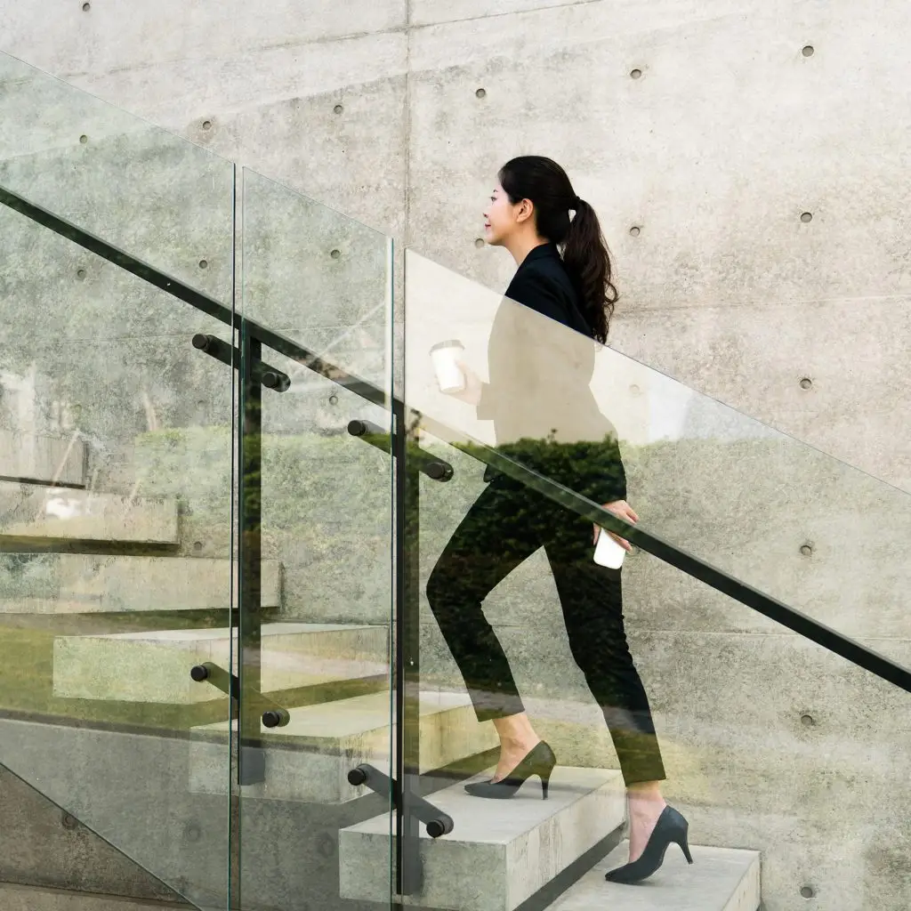 A woman in business attire confidently walks up a glass and concrete staircase, holding a coffee cup and her smartphone, exuding professionalism and purpose.