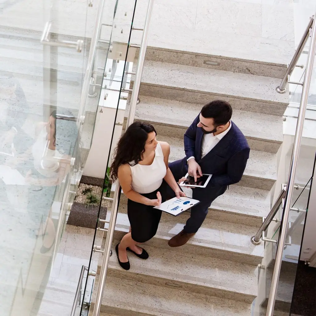 Two people in business attire stand on a staircase, discussing documents and a tablet. The scene is viewed from above, highlighting their professional conversation and collaboration on the stairs.