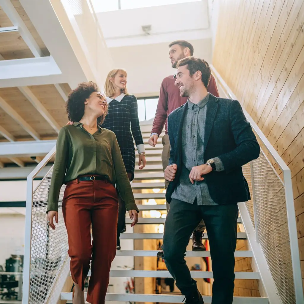 Four professionals in business attire walk down a staircase in a modern office building, conversing and sharing smiles, reflecting a positive office culture.