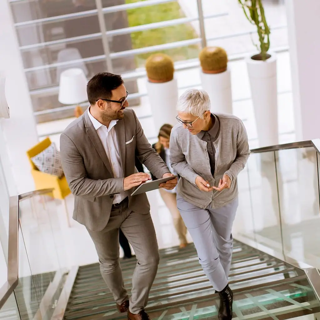 Two professionally dressed people walk up a staircase in a modern office, discussing something on a tablet, embodying teamwork and collaboration.
