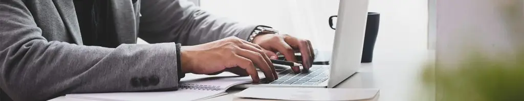 A person in a blazer types on a laptop at a desk with a notebook, pen, and coffee mug nearby.