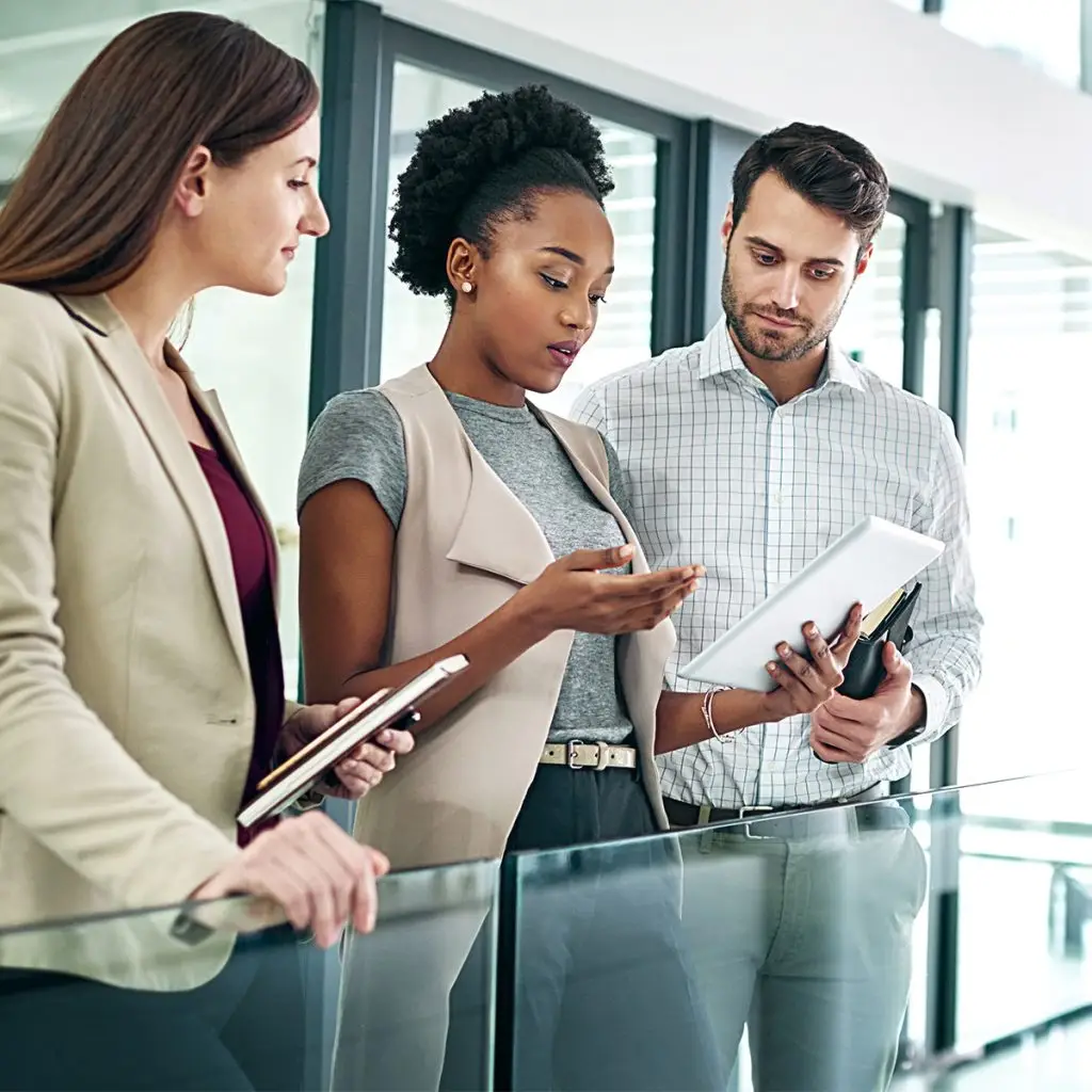 Three people in business attire stand together indoors, reviewing information on a tablet and holding notebooks, appearing to discuss work-related information.