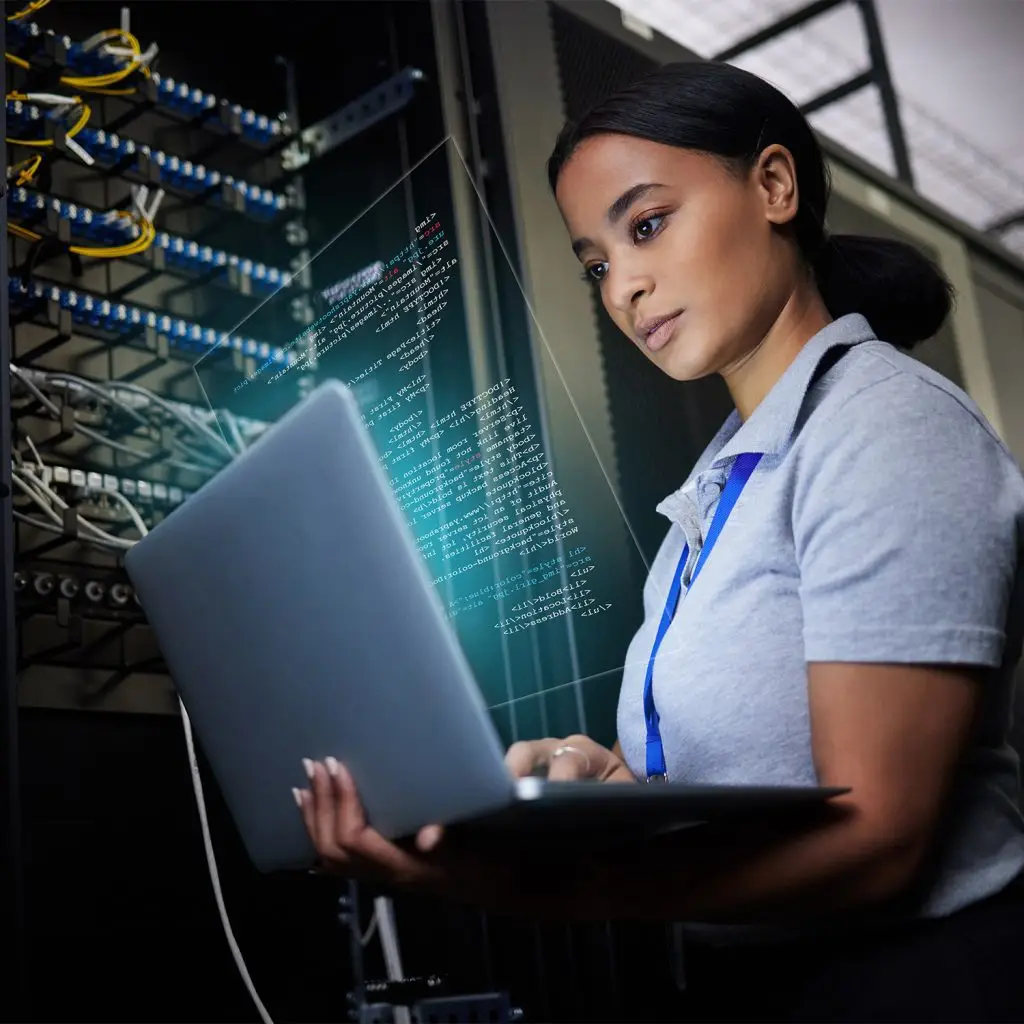 A woman stands in a server room holding a laptop, checking code displayed virtually in front of her while networking equipment is visible in the background.
