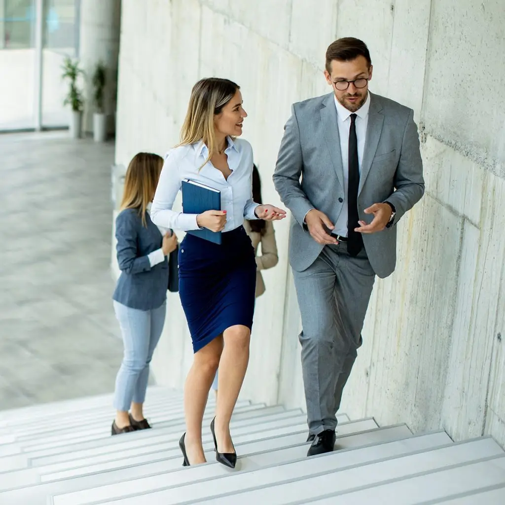 Four professionally dressed people are walking up a modern office staircase, with two in front engaged in conversation. The professional setting highlights teamwork and collaboration as they move through the building together.