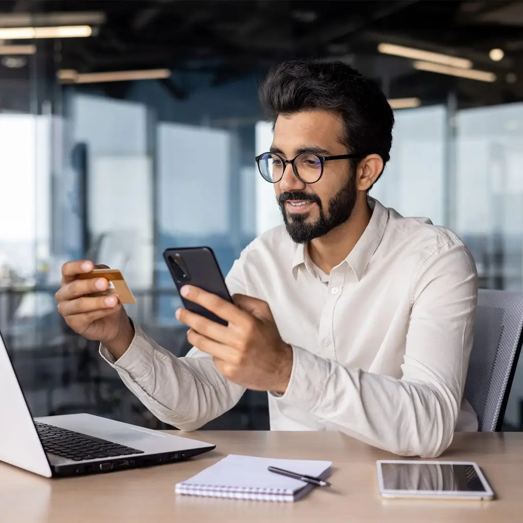 A man with glasses sits at a desk holding a credit card and smartphone, with a laptop, notepad, pen, and tablet in front of him in a modern office setting.