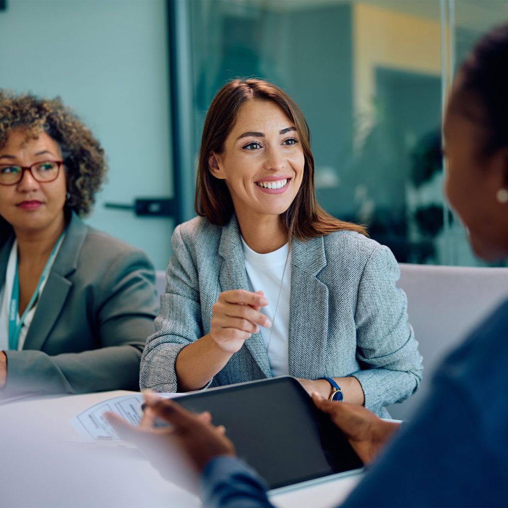 Three professionally dressed women sit at a table in an office, engaged in a discussion. One woman is smiling and gesturing while another holds a tablet, highlighting their collaborative approach to problem-solving.
