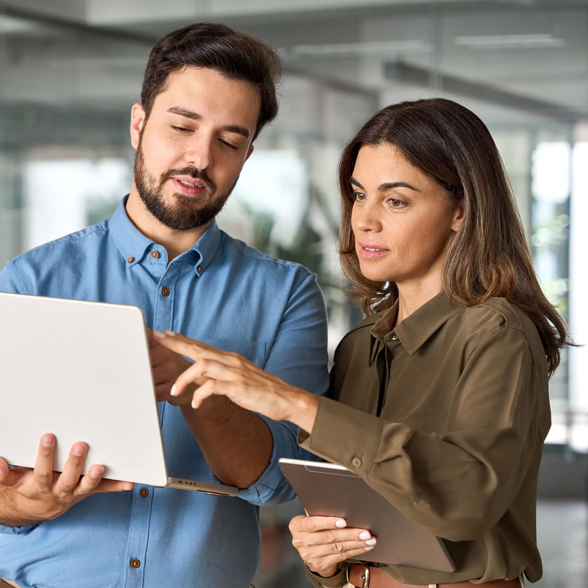 Two people standing indoors, looking at a laptop screen. One person is holding the laptop and pointing at the screen while the other holds a tablet, watching attentively as they discuss ideas.