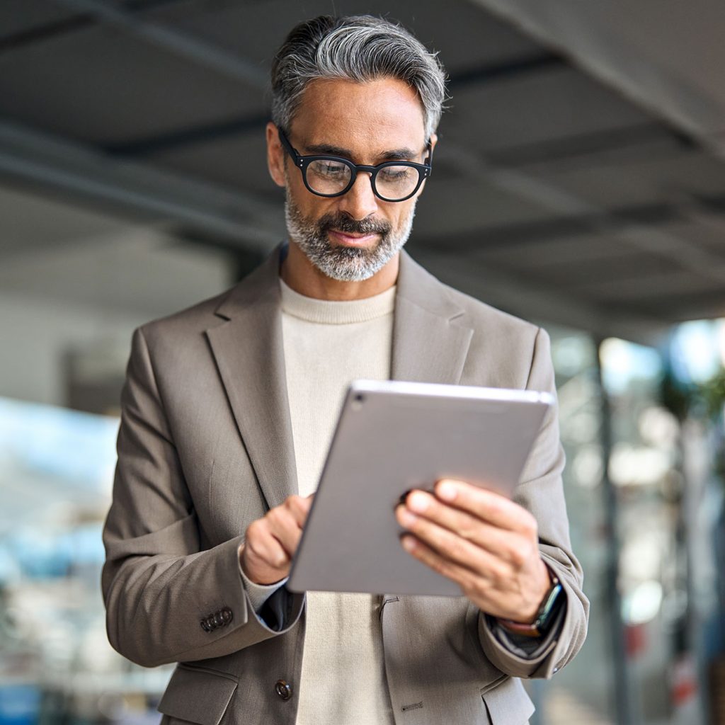 A man wearing glasses and a beige suit jacket is standing indoors, intently looking at and using a tablet device.