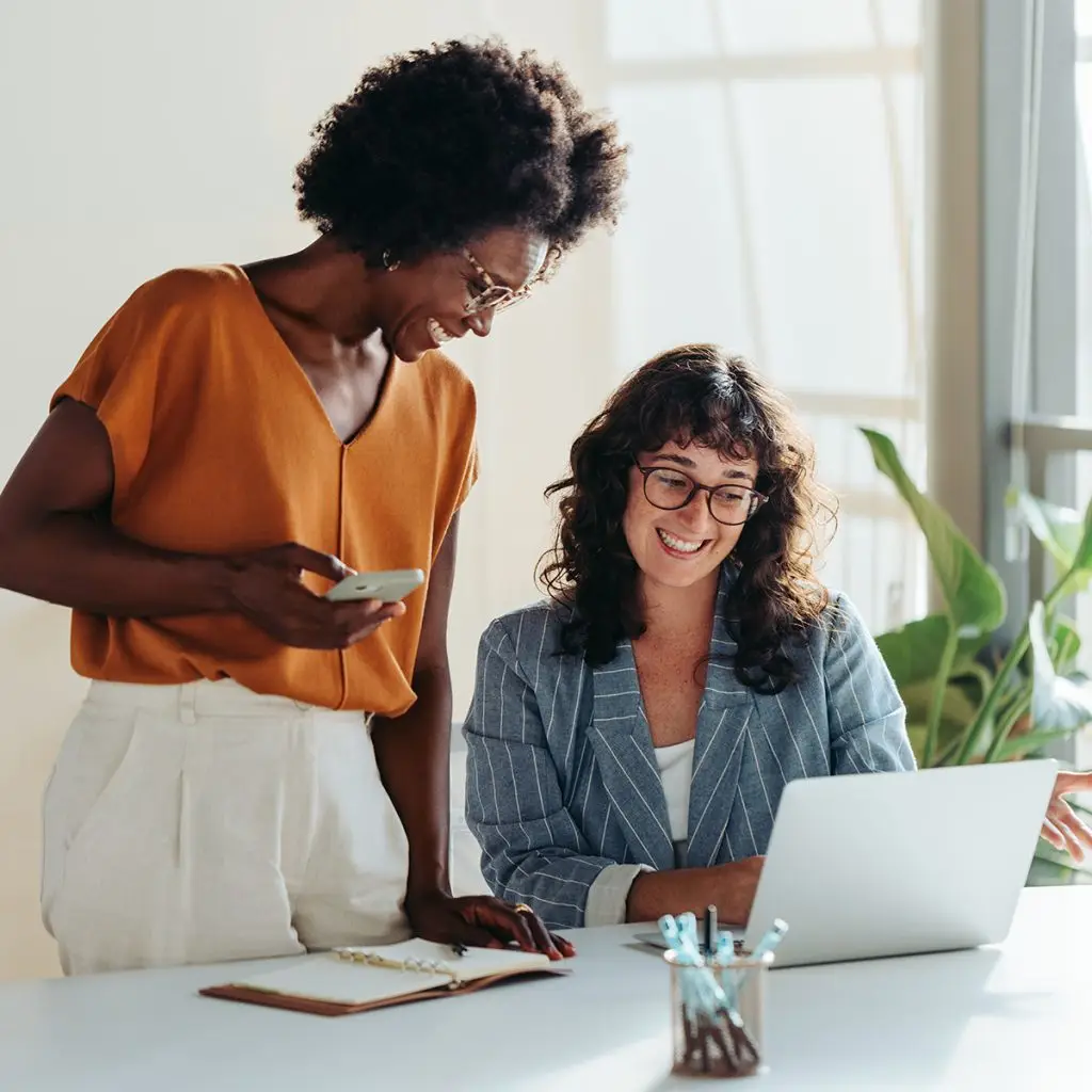 Two women collaborating at a desk, one standing with a phone and notepad, the other sitting and using a laptop, both smiling in a bright office setting that encourages teamwork and productivity.
