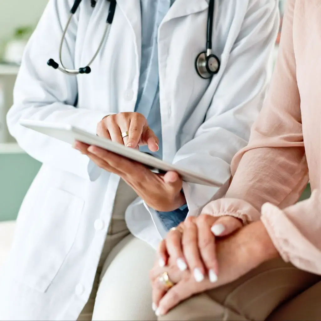 A doctor in a white coat shows HIPAA-compliant information on a tablet to a patient, who is seated and resting her hands on her lap.