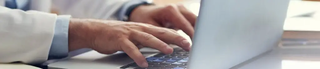 A person wearing a white coat types on a laptop keyboard at a desk, ensuring HIPAA compliance while handling sensitive information.