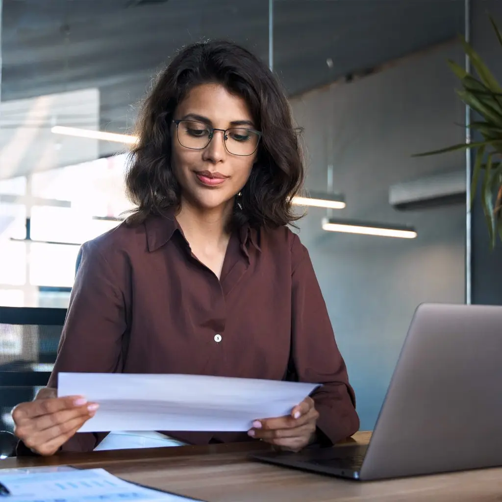 A focused woman wearing glasses sits at her office desk, reading a document intently, with her laptop and important papers arranged neatly in front of her.