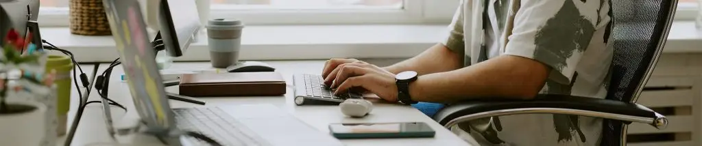 Person typing on a keyboard at a desk with a computer, smartphone, notebooks, and a reusable cup, in a well-lit office setting.