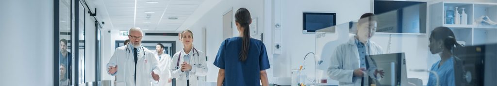 Doctors and medical staff walk and talk in a brightly lit hospital corridor with medical equipment visible through glass panels.