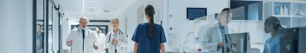 Doctors and medical staff walk and talk in a brightly lit hospital corridor with medical equipment visible through glass panels.
