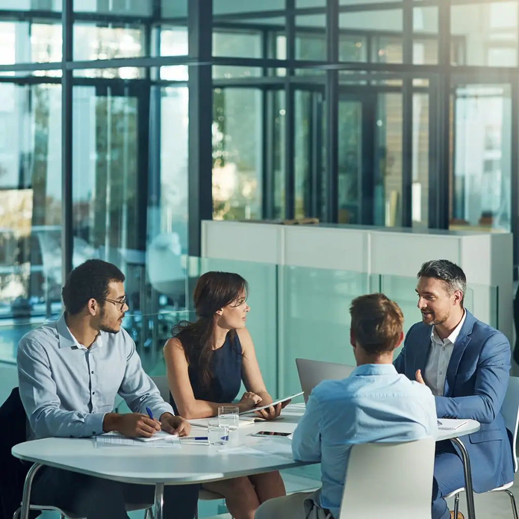 Four people in business attire sit around a table in a modern office, engaged in a discussion with laptops, papers, and glasses of water, creating a collaborative work environment.