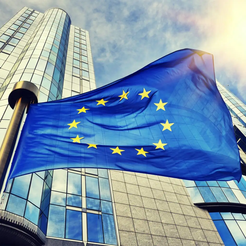 European Union flag with yellow stars on a blue background waves in front of a modern glass office building under a partly cloudy sky.