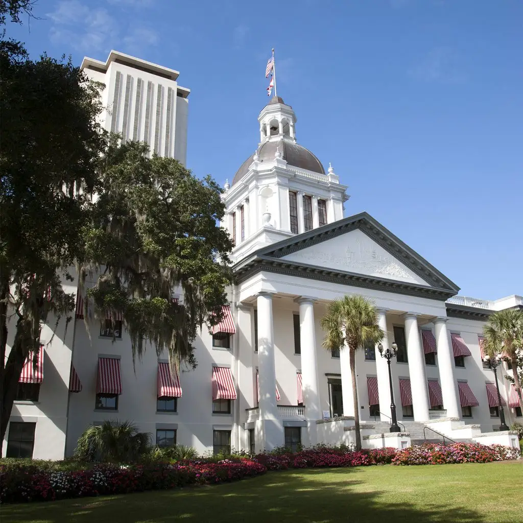 White government building with columns, dome, and red-striped awnings, surrounded by flowers and trees under a clear blue sky.