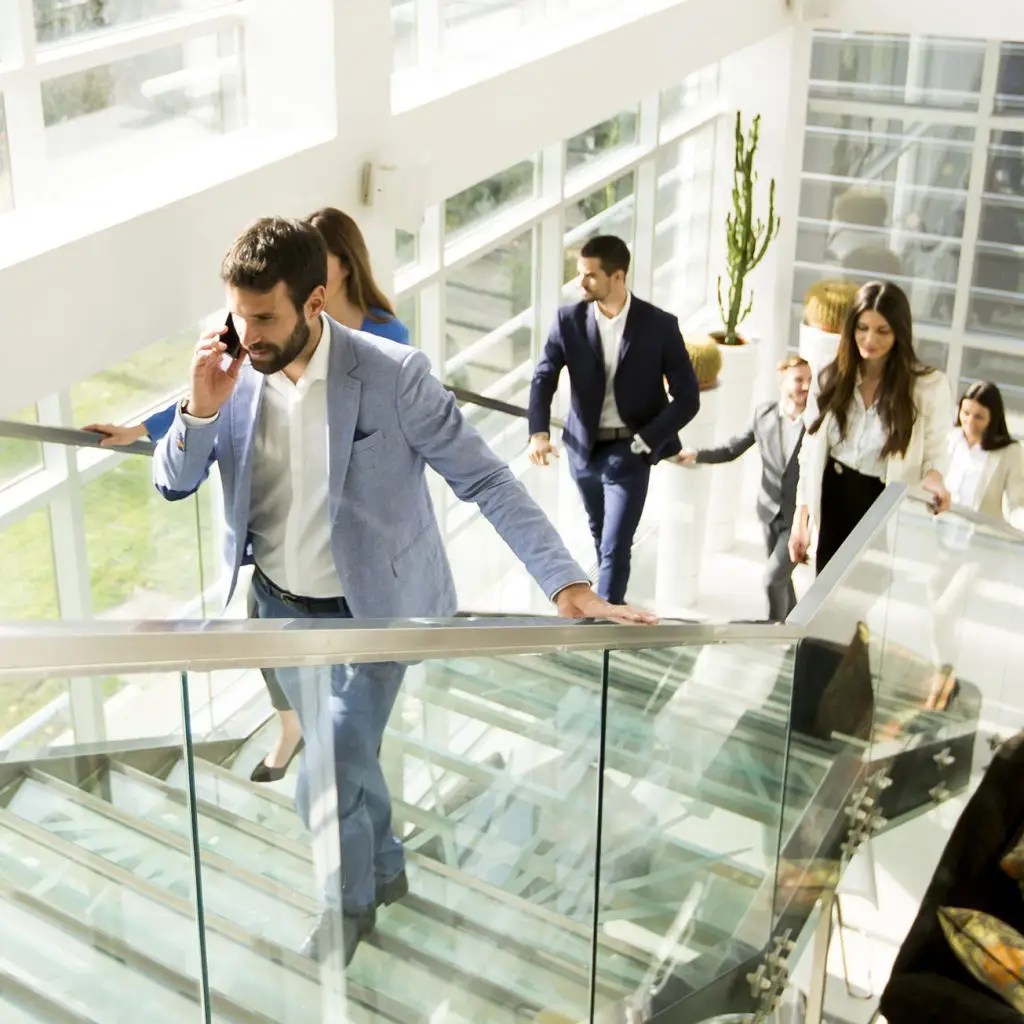 A group of professionally dressed people walk up a glass staircase in a modern office building, with one man in front talking on his phone, highlighting the dynamic pace of professional life.